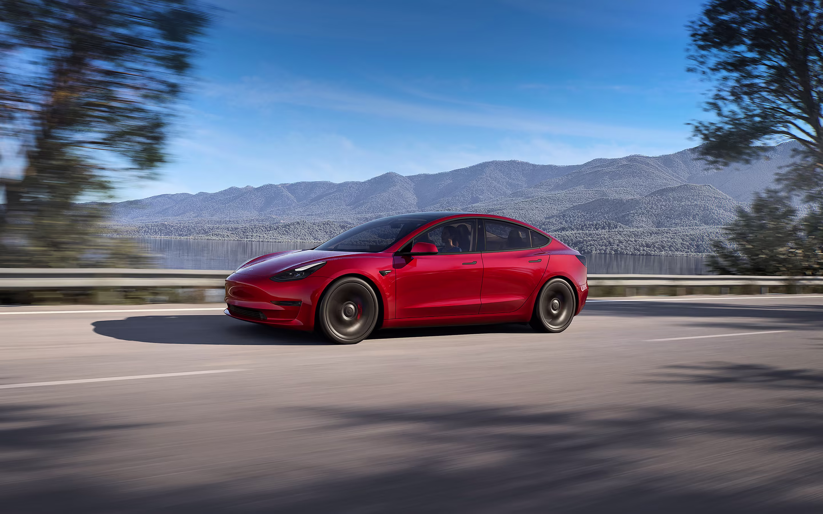 Red Model 3 driving down a lakeside road